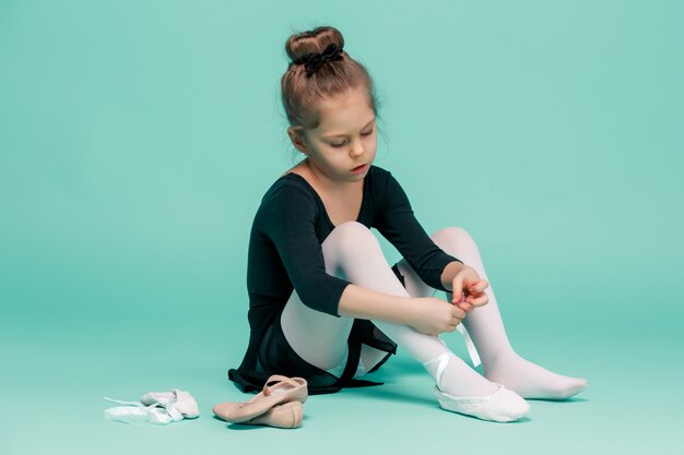 Bella piccola ballerina in abito nero per ballare seduto sul pavimento e mettere a piedi scarpe da punta su studio blu