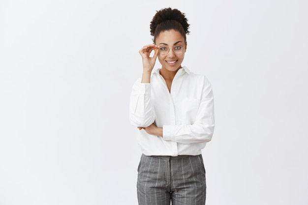 Bella psicologa elegante e intelligente con la pelle scura in camicia e pantaloni da colletto bianco, toccando gli occhiali e sorridendo con espressione sicura di sé, guardando