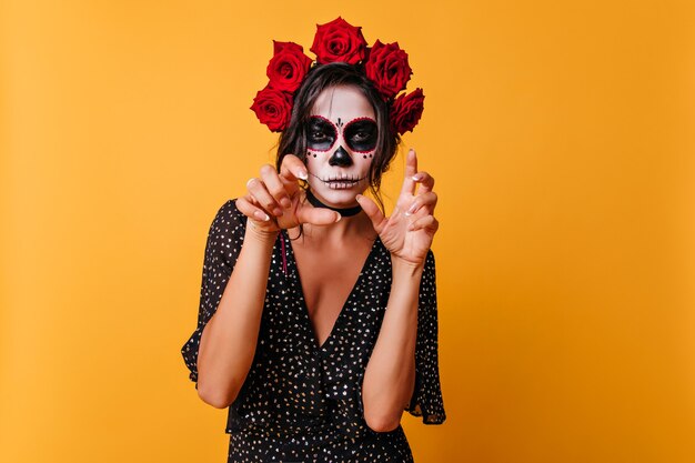 Bella ragazza abbronzata con trucco di halloween in piedi su sfondo luminoso. Meraviglioso zombie femmina con fiori nei capelli che celebra il giorno dei morti.