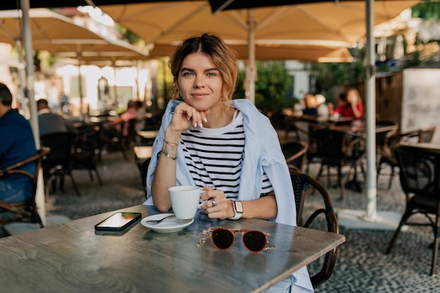 Bella ragazza affascinante con capelli mossi chiari che indossa una maglietta a righe e camicia blu è seduta su una terrazza all'aperto con caffè Bella ragazza è una ricreazione sulla terrazza estiva con caffè e smartphone