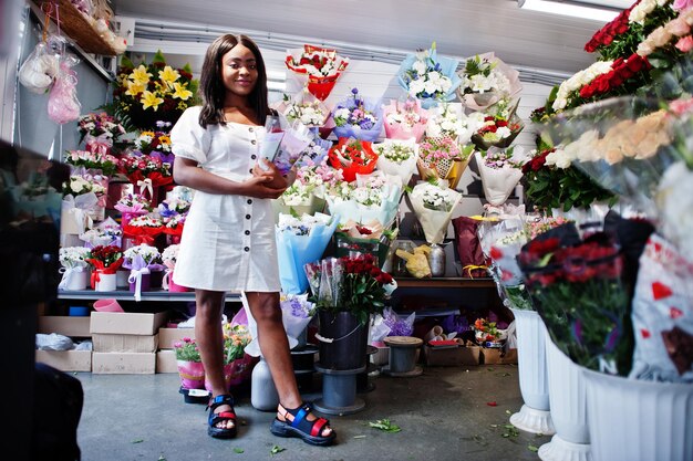 Bella ragazza afroamericana in tenero abito bianco con bouquet di fiori in mano in piedi su sfondo floreale nel negozio di fioriFioraio nero