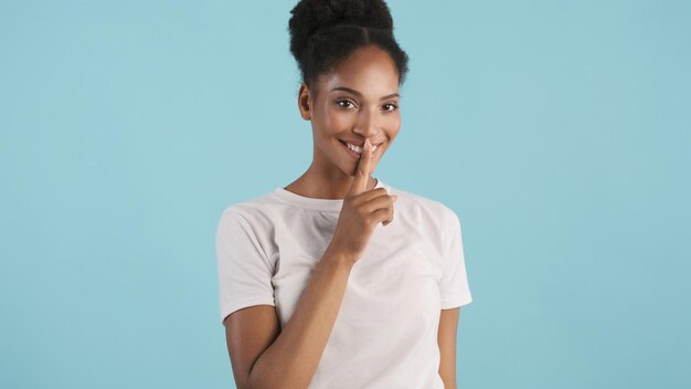 Bella ragazza afroamericana sorridente che mostra felicemente il gesto del silenzio sulla fotocamera su sfondo colorato Mantieni l'espressione segreta