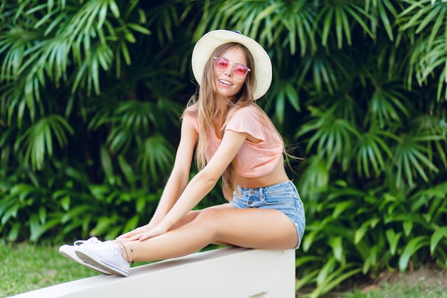 Bella ragazza alla moda che si siede sul recinto bianco nel parco tropicale con le gambe allungate.