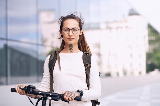 Bella ragazza alla moda con i dreadlocks sta guidando uno scooter elettrico per strada in una nuvolosa giornata estiva.