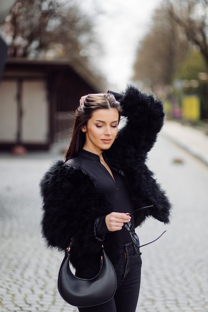 Bella ragazza alla moda dai capelli castani in vestito nero all'aperto. Ritratto di giovane donna elegante attraente con capelli lunghi in primavera nelle strade della città.