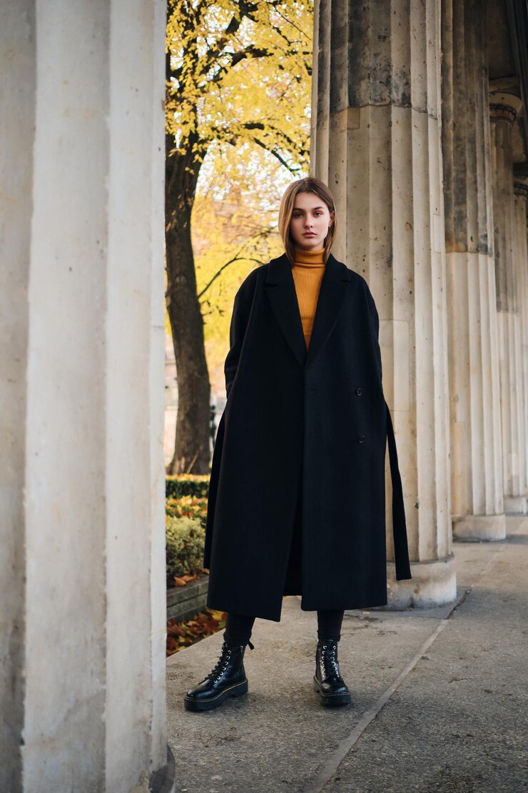 Foto gratuito bella ragazza alla moda in cappotto nero e stivali che guardano intensamente nella fotocamera all'aperto