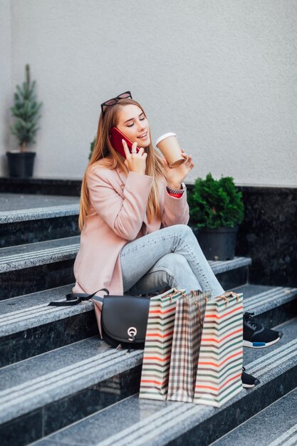 Bella ragazza alla moda seduta per strada, dopo lo shopping, tenendo il caffè sulle mani, in posa all'aperto.