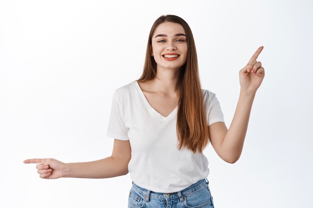 Bella ragazza allegra, sorridente e danzante, puntando il dito lateralmente su due offerte promozionali, mostrando le scelte di banner, le migliori offerte promozionali, in piedi sul muro bianco