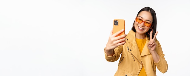 Bella ragazza asiatica alla moda in occhiali da sole che prende selfie sullo smartphone che posa per la foto che tiene il fondo bianco dello studio del telefono cellulare