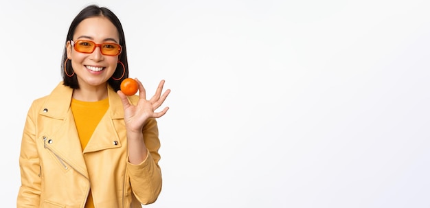 Bella ragazza asiatica in occhiali da sole che mostrano mandarino e sorridente che sembra felice in posa in giallo su sfondo per studio
