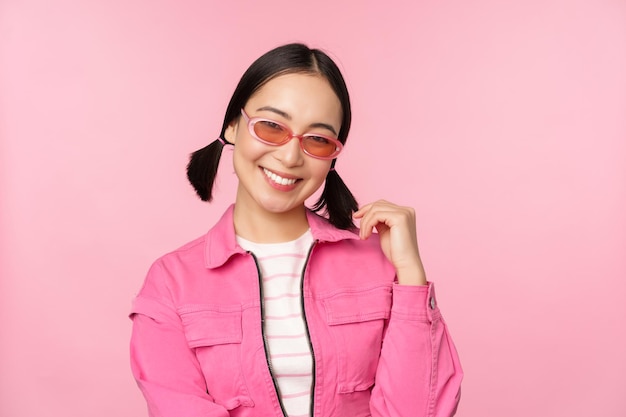 Bella ragazza asiatica in sunlgasses che sorride alla macchina fotografica che posa contro il fondo rosa dello studio