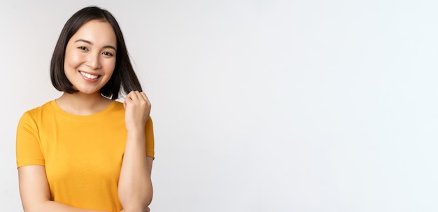 Bella ragazza asiatica romantica che sorride e che gioca con i capelli che guardano felice alla macchina fotografica che sta in maglietta gialla sopra priorità bassa bianca