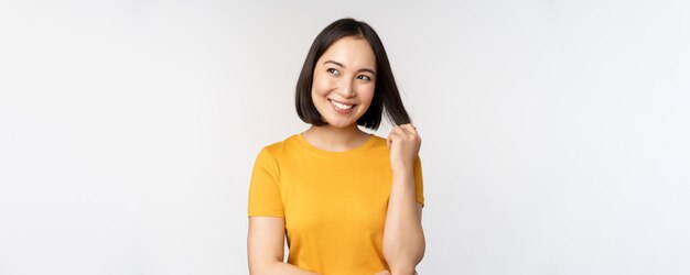 Bella ragazza asiatica romantica che sorride e che gioca con i capelli che guardano felice alla macchina fotografica che sta in maglietta gialla sopra priorità bassa bianca