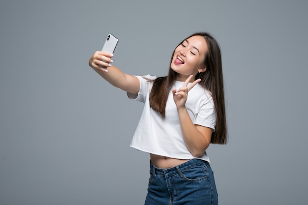 Bella ragazza asiatica socievole che prende selfie o che parla sulla video chiamata facendo uso del telefono cellulare sopra fondo grigio