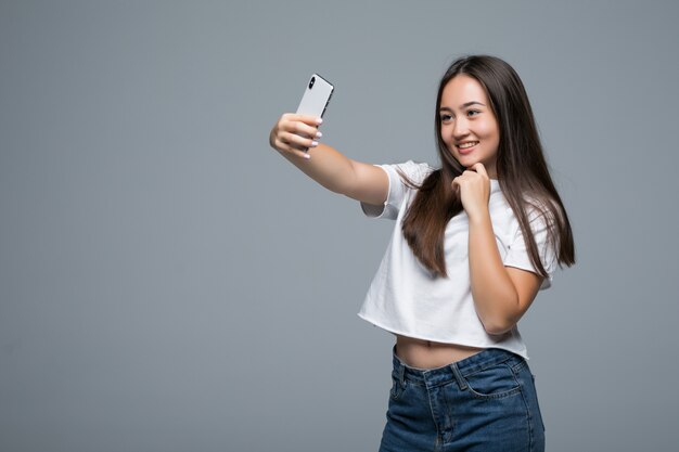 Bella ragazza asiatica socievole che prende selfie o che parla sulla video chiamata facendo uso del telefono cellulare sopra fondo grigio