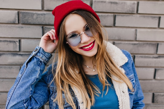 Bella ragazza bianca con un sorriso allegro divertendosi nella fredda giornata di primavera. Outdoor ritratto di gioiosa donna bionda indossa occhiali blu e cappello rosso.