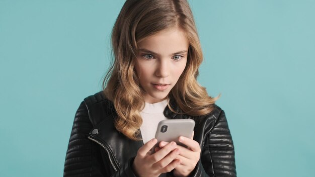 Bella ragazza bionda adolescente che sembra eccitata pensando a come rispondere al messaggio su sfondo blu. Tecnologia moderna