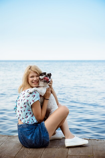 Bella ragazza bionda che si siede, giocando con il bulldog francese vicino al mare