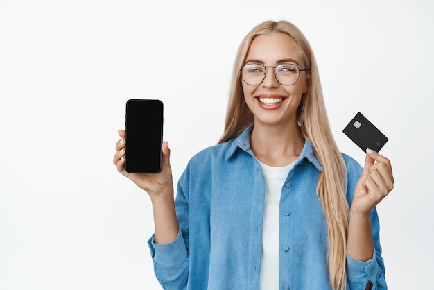 Bella ragazza bionda con gli occhiali sorridente che mostra lo schermo del telefono cellulare e la carta di credito che dimostra l'app finanziaria in piedi su sfondo bianco