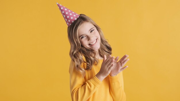 Bella ragazza bionda eccitata che indossa il cappello di compleanno che sembra allegro su sfondo giallo Ragazza teenager felice di vedere gli amici