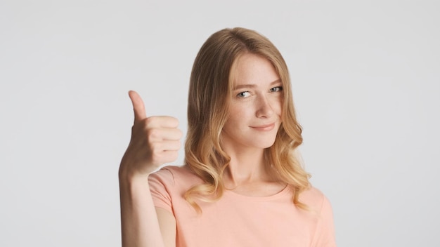 Bella ragazza bionda sicura mantenendo il pollice sulla fotocamera isolata su sfondo bianco Concetto di linguaggio del corpo