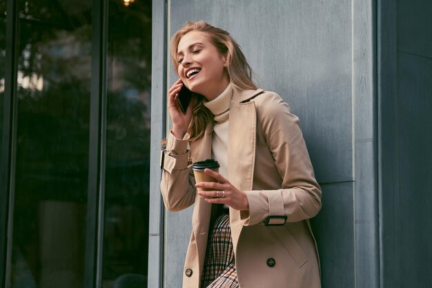 Bella ragazza bionda sorridente in trench elegante con caffè per andare a parlare felicemente sul cellulare all'aperto