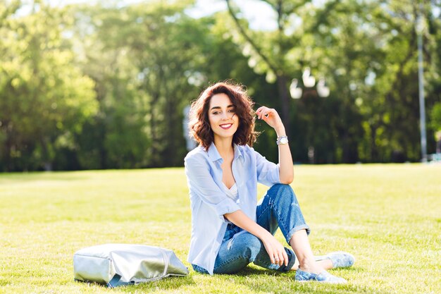 Bella ragazza bruna con i capelli corti in posa sull'erba nel parco. Indossa maglietta bianca, camicia e jeans, scarpe. Sembra felice alla luce del sole.