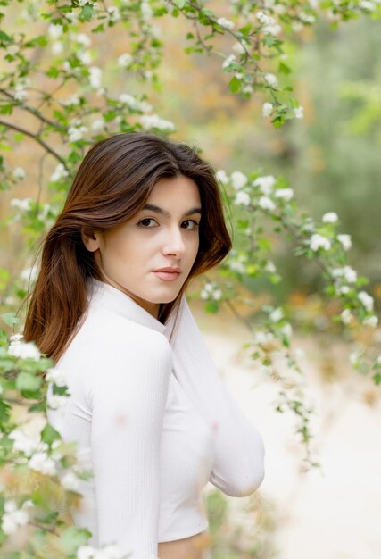 Bella ragazza bruna con servizio fotografico in piedi nell'albero in fiore che guarda l'obbiettivo