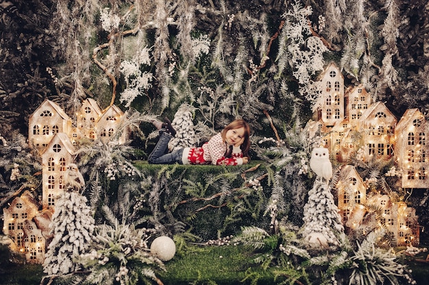 Bella ragazza bruna in maglione invernale e jeans posa su prato verde circondato da case fatte a mano illuminate e rami di abete ricoperti di neve artificiale. Fiaba di Natale.