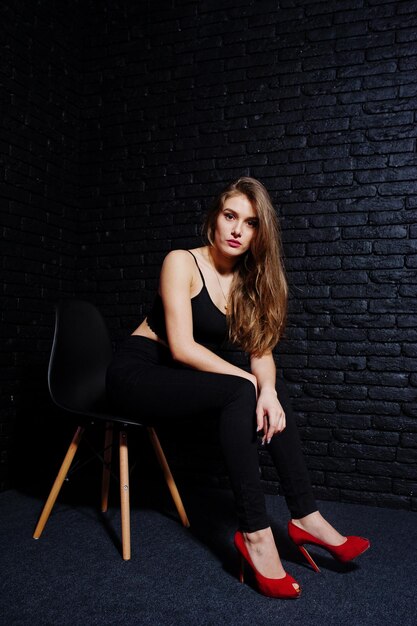 Bella ragazza bruna indossa su tacchi alti neri e rossi seduta e posa su sedia in studio contro muro di mattoni scuri Ritratto del modello in studio