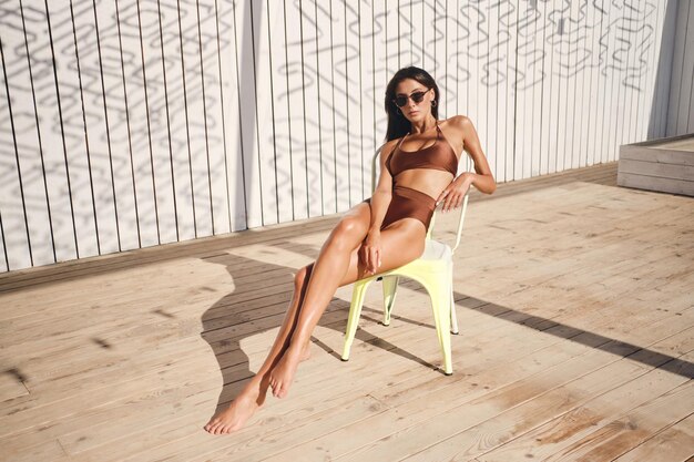 Bella ragazza bruna sexy in costume da bagno e occhiali da sole guardando premurosamente a porte chiuse a prendere il sole sulla sedia sulla spiaggia