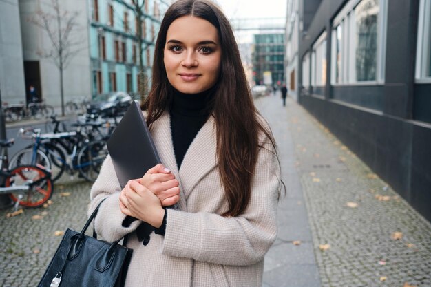 Bella ragazza bruna sorridente con il computer portatile che guarda felicemente a porte chiuse sulla strada della città