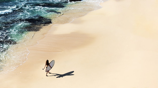 Bella ragazza cammina con una tavola da surf su una spiaggia selvaggia. splendida vista dall'alto.