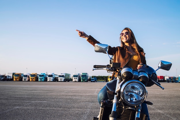 Bella ragazza castana che si siede sulla motocicletta di stile retrò e punta il dito verso l'alto