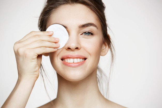 Bella ragazza castana con l'occhio nascondentesi pulito della pelle perfetta dietro la spugna di cotone che sorride esaminando macchina fotografica sopra fondo bianco. Cosmetologia e spa.