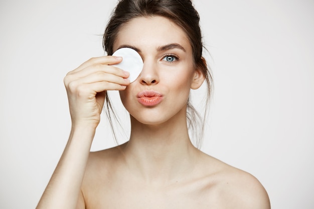 Bella ragazza castana con l'occhio nascondentesi pulito della pelle perfetta dietro la spugna di cotone che sorride esaminando macchina fotografica sopra fondo bianco. Cosmetologia e spa.