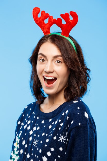 Bella ragazza castana sorpresa giovani in corna tricottate della renna di natale e del maglione che sorride sopra la parete blu