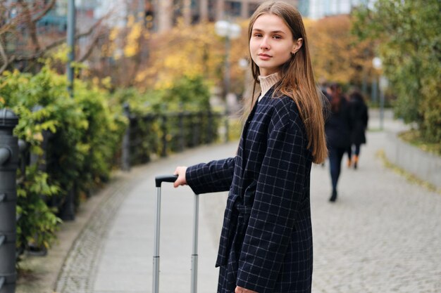 Bella ragazza casual in cappotto che guarda sognante a porte chiuse camminando per strada con la valigia