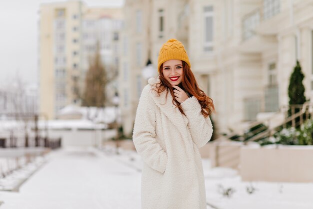 Bella ragazza caucasica che cammina intorno alla città nella giornata invernale. Donna felice dello zenzero in camice bianco in posa sulla strada.