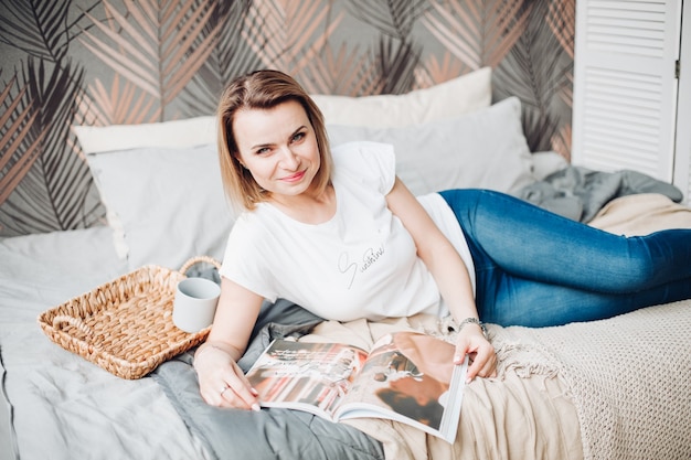 Bella ragazza caucasica con capelli biondi, maglietta bianca, jeans si trova nella grande camera da letto luminosa e legge una rivista