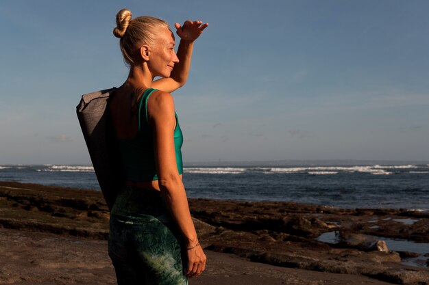 Bella ragazza che cammina sulla spiaggia con il tappetino da yoga