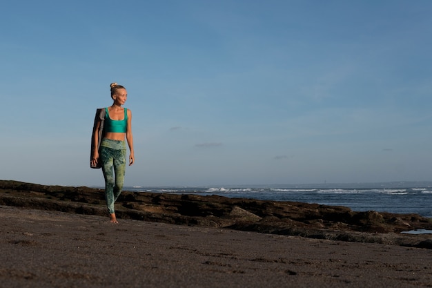 Bella ragazza che cammina sulla spiaggia con il tappetino da yoga