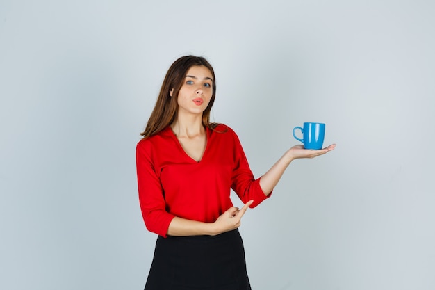 Bella ragazza che mostra tazza in camicetta rossa, gonna nera e sembra attraente, vista frontale.