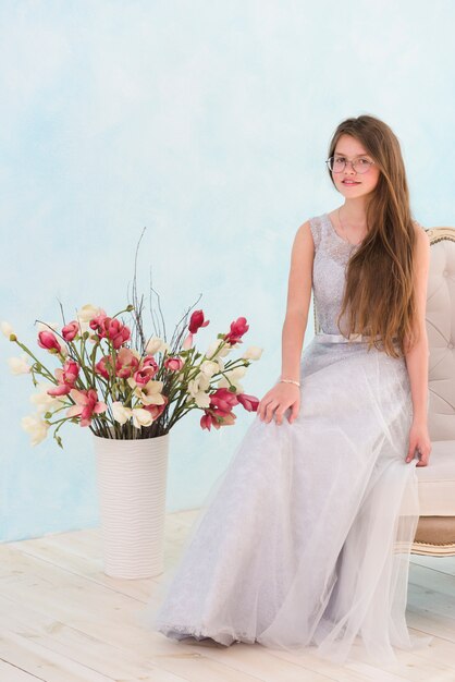 Bella ragazza che si siede sulla poltrona vicino al vaso di fiori che guarda l&#39;obbiettivo