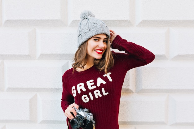 Bella ragazza con capelli lunghi e labbra rosse in maglione marsala sul muro grigio. Indossa un cappello lavorato a maglia, tiene le mani di camer ain e sorride.