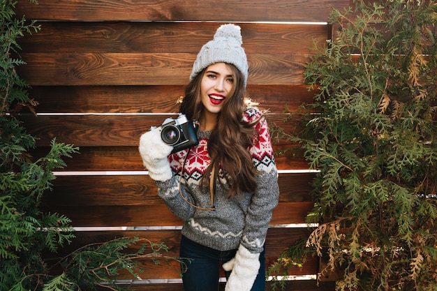 Bella ragazza con capelli lunghi in cappello lavorato a maglia e guanti bianchi su rami verdi circondano in legno. Indossa un maglione caldo, tiene la macchina fotografica, sembra goduta.