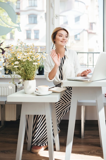 Bella ragazza con i capelli biondi che si siede nel ristorante con il computer portatile, una tazza di caffè e un mazzo di camomilla sul tavolo. Ritratto di ragazza sorridente che guarda da parte sventolando e salutando al caffè.