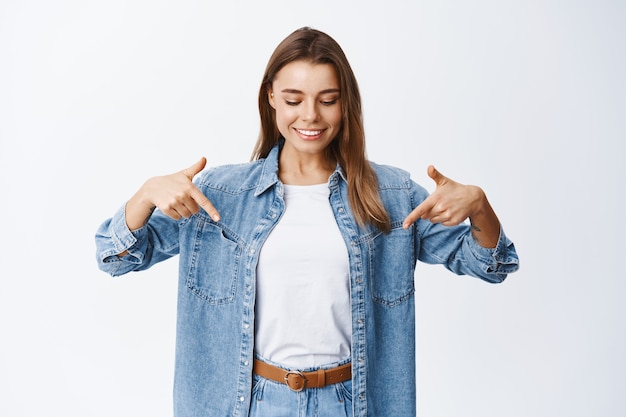 Bella ragazza con i capelli biondi e un sorriso bianco che punta le dita verso il basso, guardando lo spazio della copia in basso, mostrando il tuo logo qui, parete dello studio