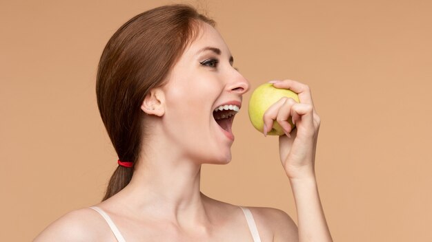Bella ragazza con i capelli legati sulla schiena che mangia una mela gustosa a pranzo. Vista laterale del modello attraente che promuove uno stile di vita sano. Donna castana con i denti bianchi che tengono in mano deliziosi frutti.