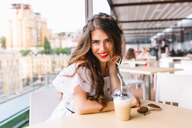 Bella ragazza con i capelli lunghi è seduta al tavolo sulla terrazza del caffè. Indossa un abito bianco con spalle nude e rossetto rosso. Sta sorridendo alla telecamera.